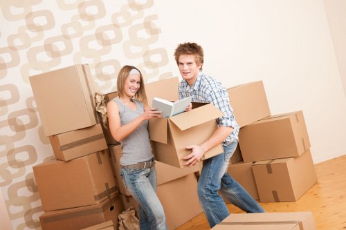 A young man and woman are inside a room with a cream-colored wall featuring a geometric pattern, surrounded by multiple large cardboard moving boxes of various sizes, some stacked on the floor and others being carried. The woman, standing to the left, has shoulder-length blonde hair and is wearing a sleeveless grey top and denim jeans; she is smiling and holding a tablet device. The young man, standing to the right, has short, light brown hair and is dressed in a blue and white checkered shirt with matching jeans; he is in the process of carrying a medium-sized cardboard box. The scene appears to be part of a house removal operation, with the boxes and relaxed expressions indicating packing or unpacking activity typical of a relocation service in Roehampton, London, or nearby areas. The room has wooden flooring and is well-lit, suggesting natural light from nearby windows, consistent with an interior space prepared for moving or storage purposes.