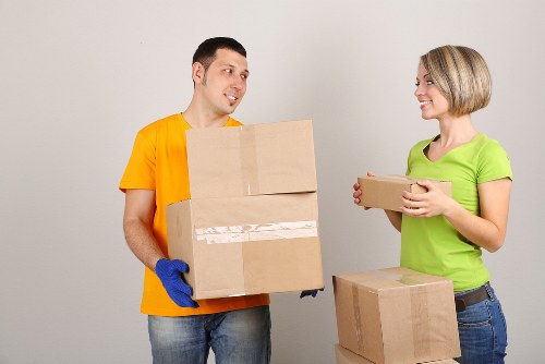 A male and female mover are indoors in a residential property, involved in packing or unpacking during a house removal process. The male, wearing a bright yellow T-shirt and blue gloves, is holding two cardboard boxes stacked together, with a slight smile. The female, dressed in a lime green T-shirt and jeans, is holding a smaller cardboard box, smiling as she faces the male. There are additional cardboard boxes on the floor nearby, some stacked, indicating an active move. The background features a plain, light-colored wall, suggesting a modern interior setting. The lighting is bright and even, highlighting the textures of the cardboard boxes and casual clothing. This scene reflects the typical activities involved in house removals in Roehampton or nearby areas, emphasizing careful handling of household items during a relocation.