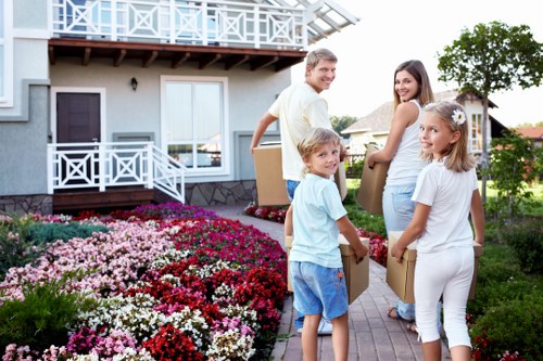 A family of four, consisting of a man, a woman, a young boy, and a young girl, are standing outside a light grey two-storey house with white window frames and a wooden balcony in a residential neighbourhood, preparing to move. The man, dressed in a white shirt and beige trousers, is holding a cardboard box and smiling at the camera. The woman, wearing a light blue blouse and white trousers, is also holding a box and facing the man, smiling. The children, a boy in a light blue shirt and denim shorts, and a girl in white attire, are holding smaller boxes and looking towards the camera, appearing eager. The front garden is neatly landscaped with vibrant pink, red, and white flowers, and a paved walkway leads from the sidewalk to the house. The background includes trees and other houses, indicating a suburban setting. This scene illustrates a typical residential move with a family engaging in house removals, supported by Roehampton Man with Van's services, in the Roehampton area near SW15 postcode.