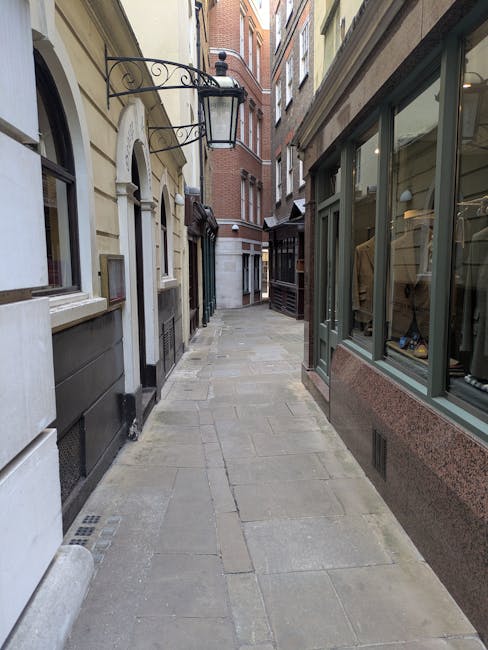 A narrow pedestrian street with a paved stone surface, flanked by historic buildings with a mix of brick and stone facades. On the right, there is a shop window displaying clothing and mannequins insi